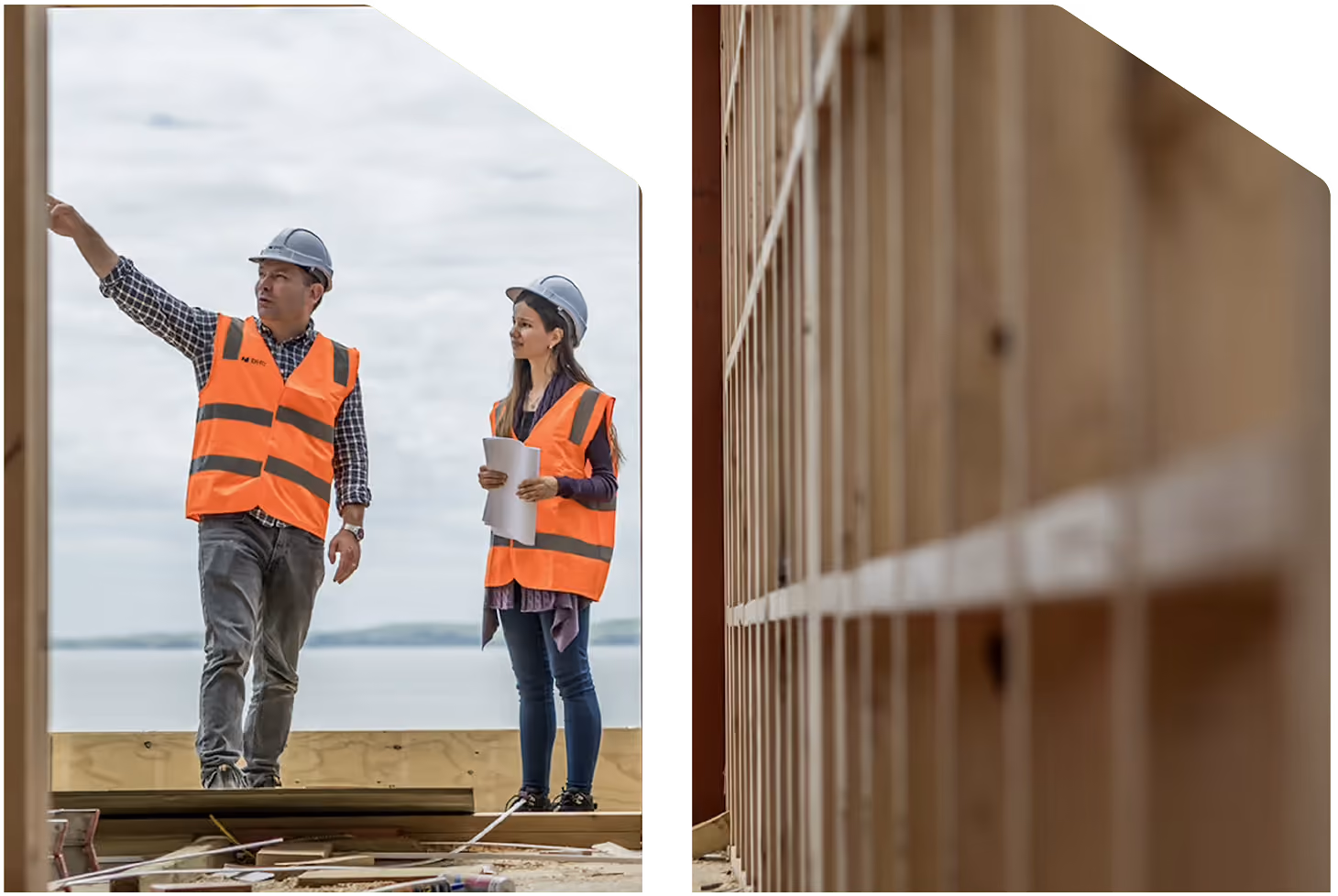 Two construction workers wearing orange safety vests and helmets stand on a construction site near wooden framing; one points ahead while the other holds blueprints, with an ocean in the background.