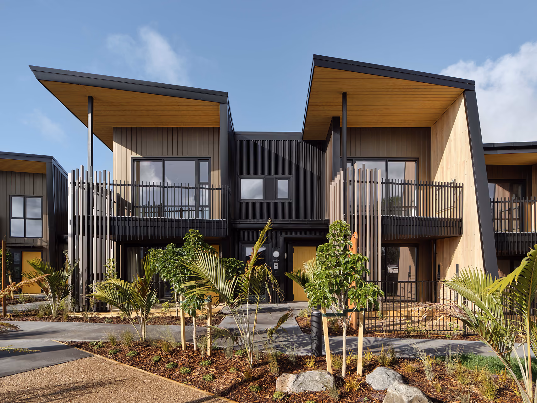 Modern two-story residential building with angled roofs and a mix of wood and black siding, featuring balconies and landscaped front with small trees and plants.