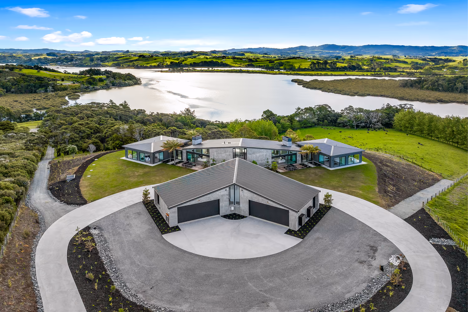 A modern house with a unique angular design surrounded by a circular driveway, situated on a green hill overlooking a large body of water and distant rolling hills under a blue sky with scattered clouds.