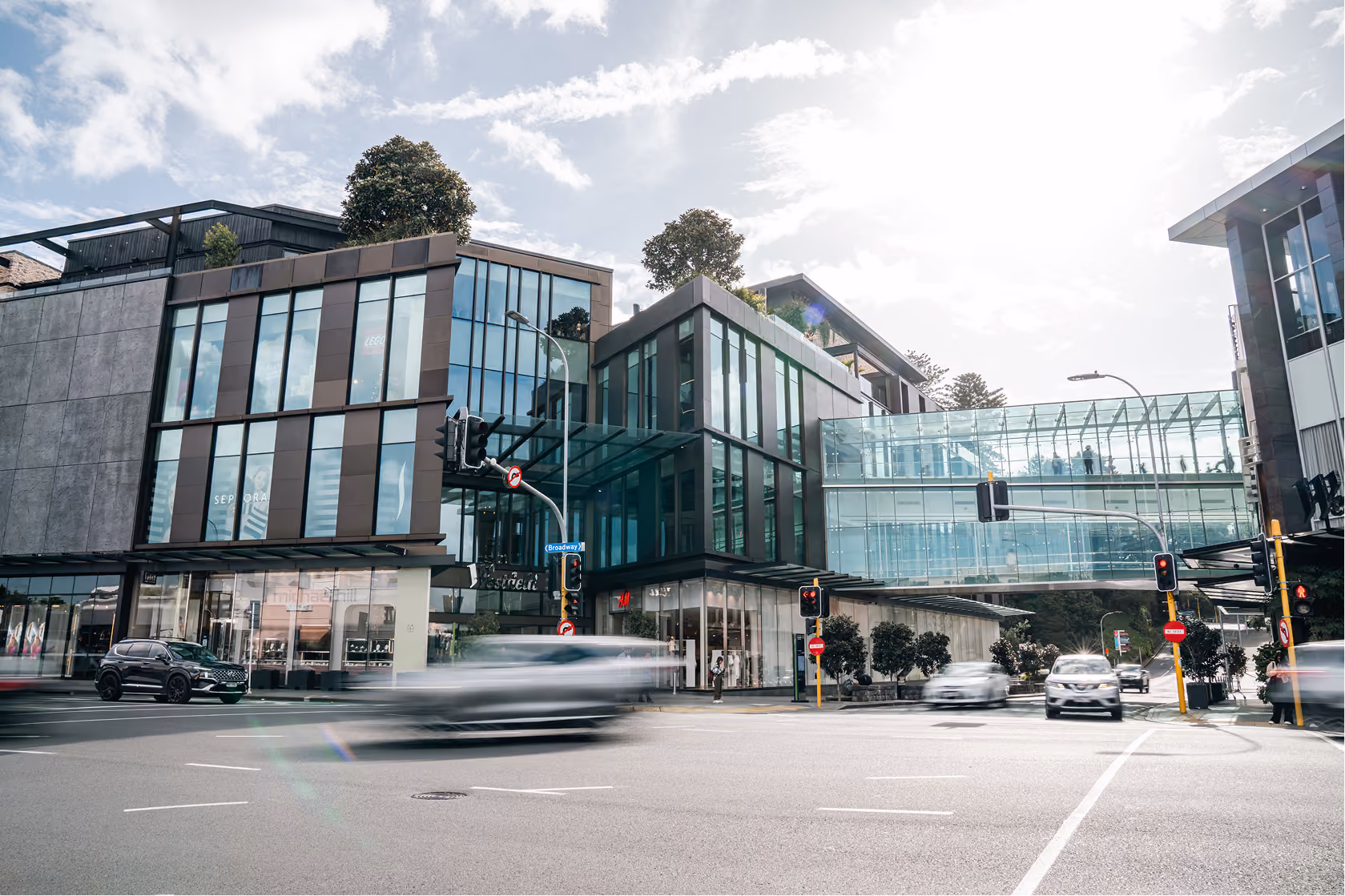 Modern glass and concrete buildings with a pedestrian bridge above a busy urban intersection with traffic and blurred moving cars under a cloudy sky.