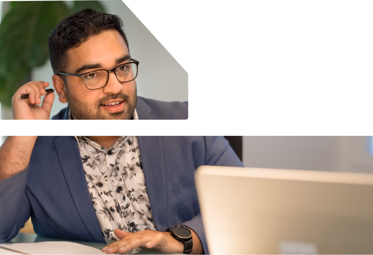 Man with glasses and a beard wearing a blue blazer and floral shirt, holding a pen near his ear while working at a laptop.