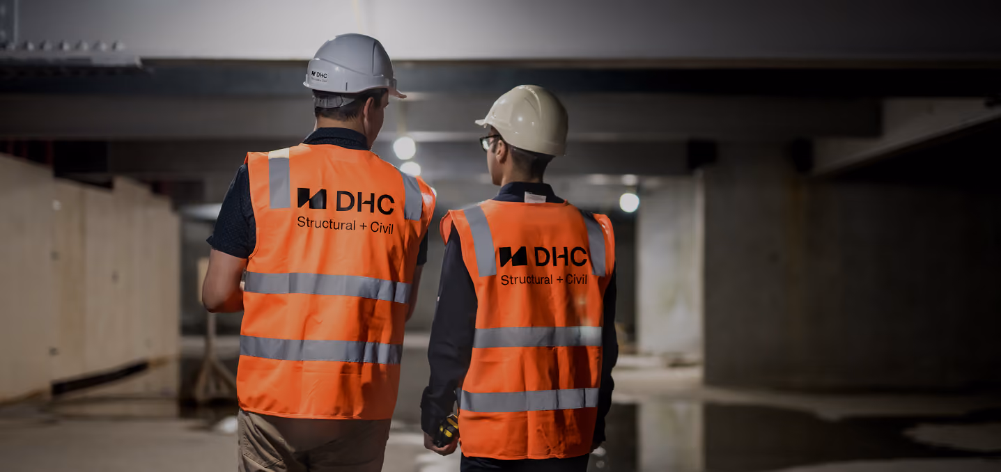 Two construction workers wearing white helmets and orange reflective vests with DHC Structural + Civil logos walking inside a dimly lit building under construction.