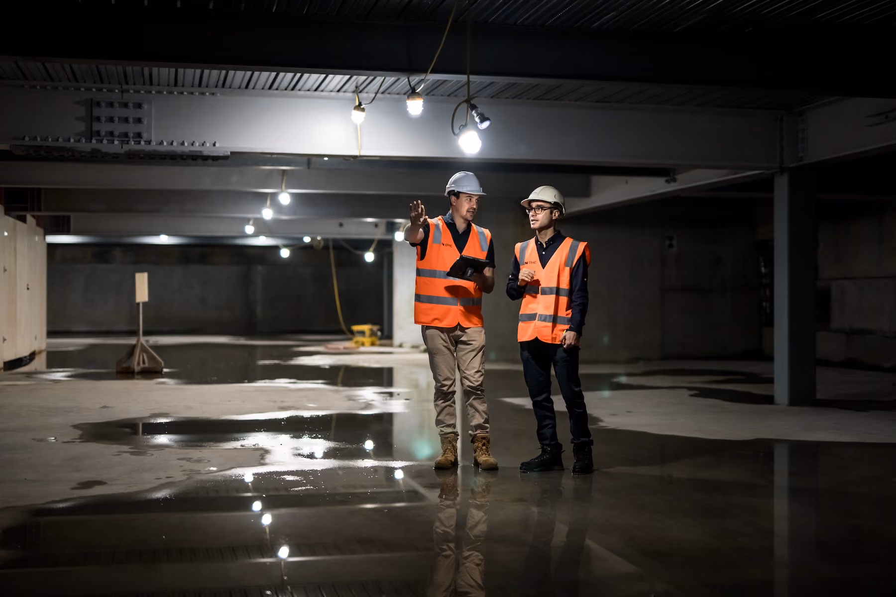 Two construction workers in hard hats and orange safety vests standing and talking inside a dimly lit building with a wet concrete floor reflecting the overhead lights.