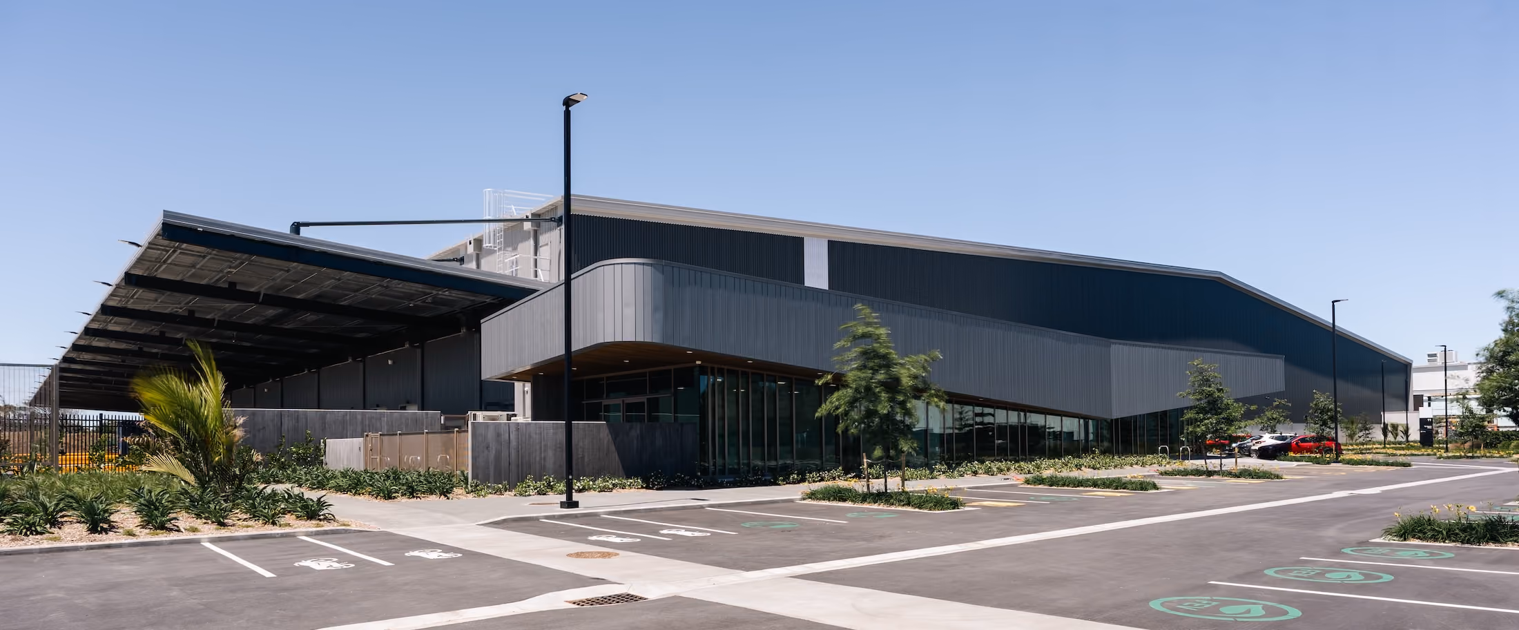 Modern industrial warehouse building with black and gray exterior, surrounded by parking spaces and small landscaped areas with trees and plants, under a clear blue sky.