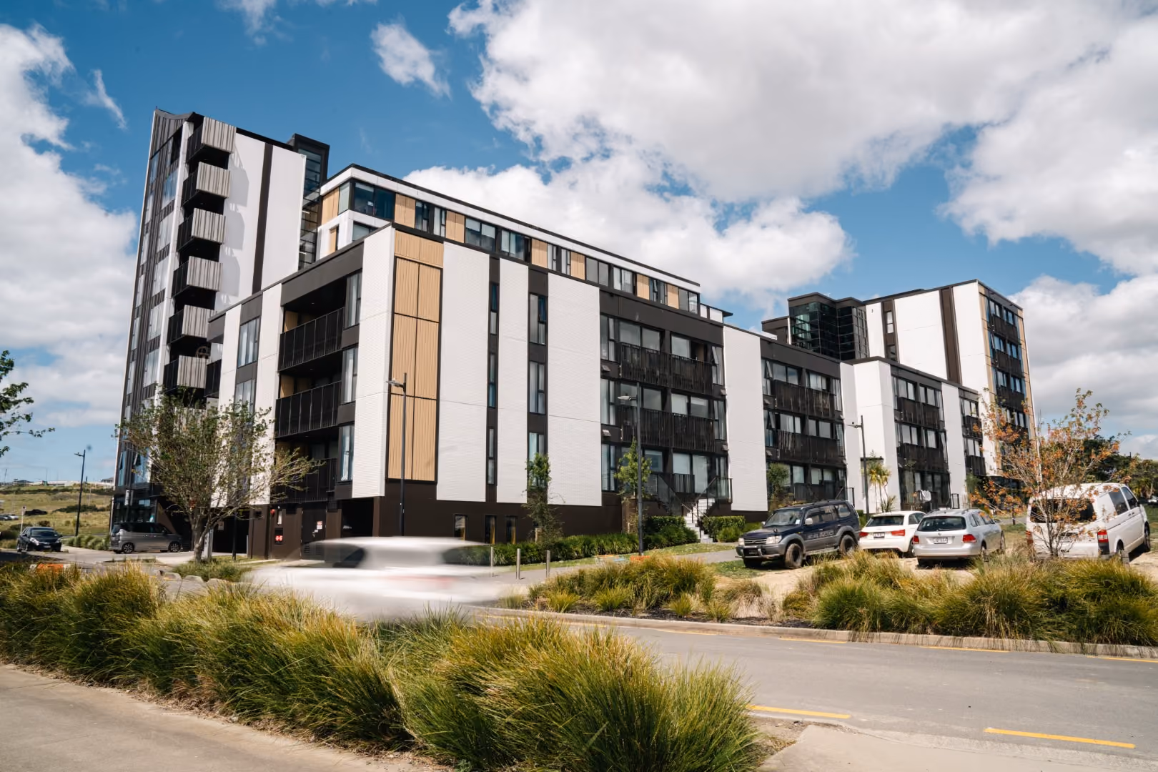 Modern multi-story residential building with white, black, and wood-paneled exterior under a partly cloudy blue sky, surrounded by greenery and parked cars on a nearby street.