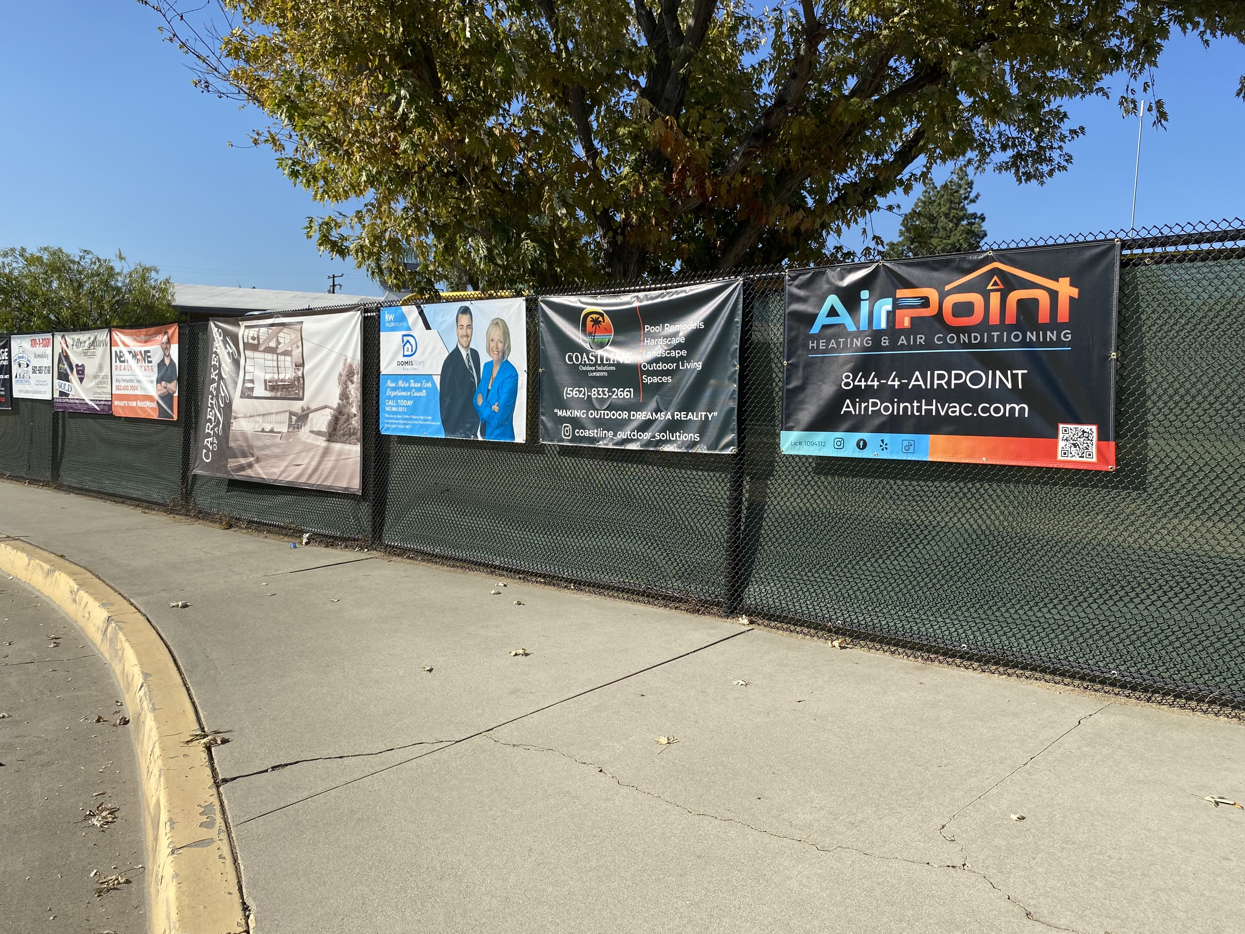Advertising banners on a chain-link fence with a tree and blue sky
