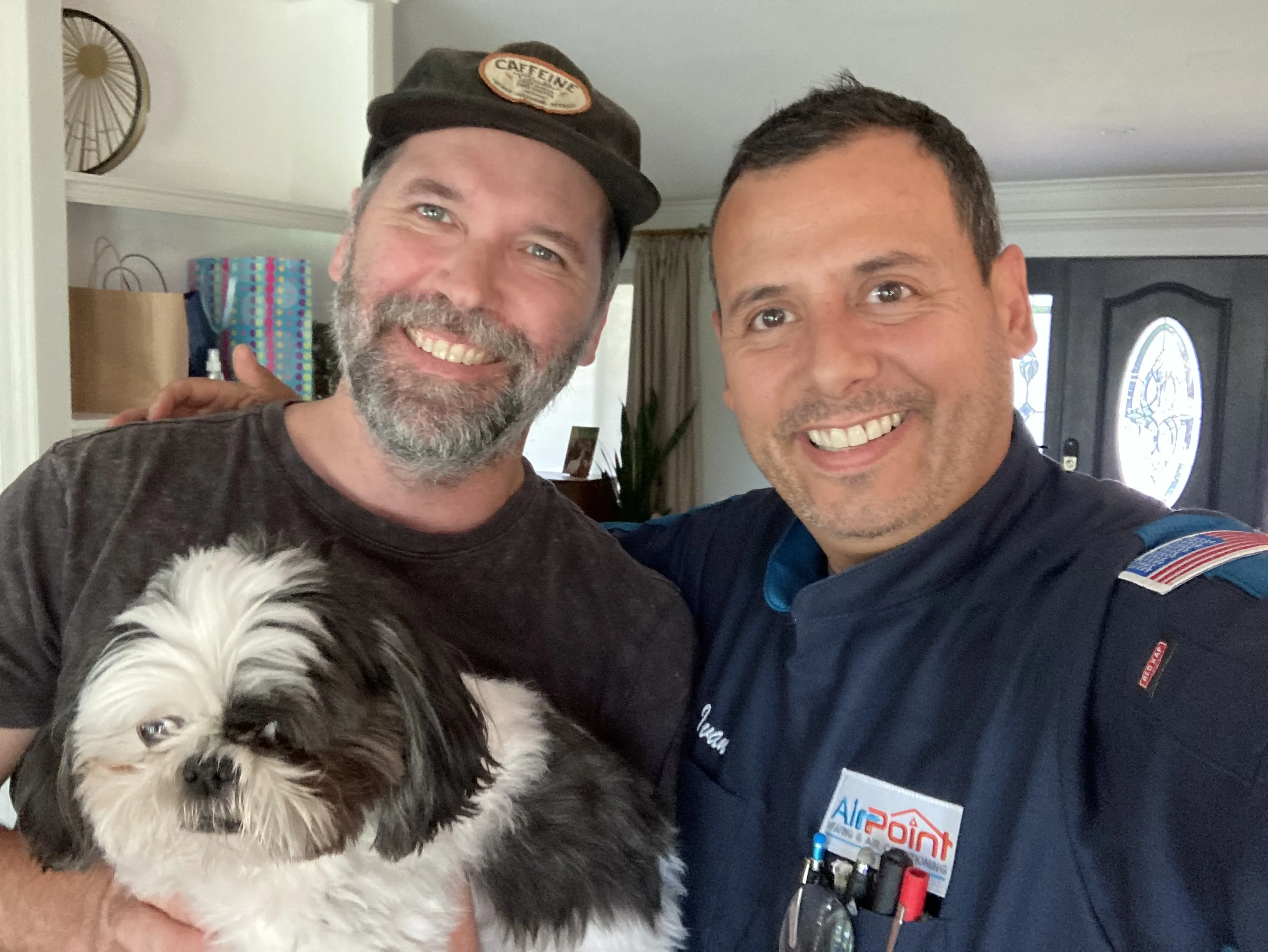 Two smiling men with a black and white dog inside a home