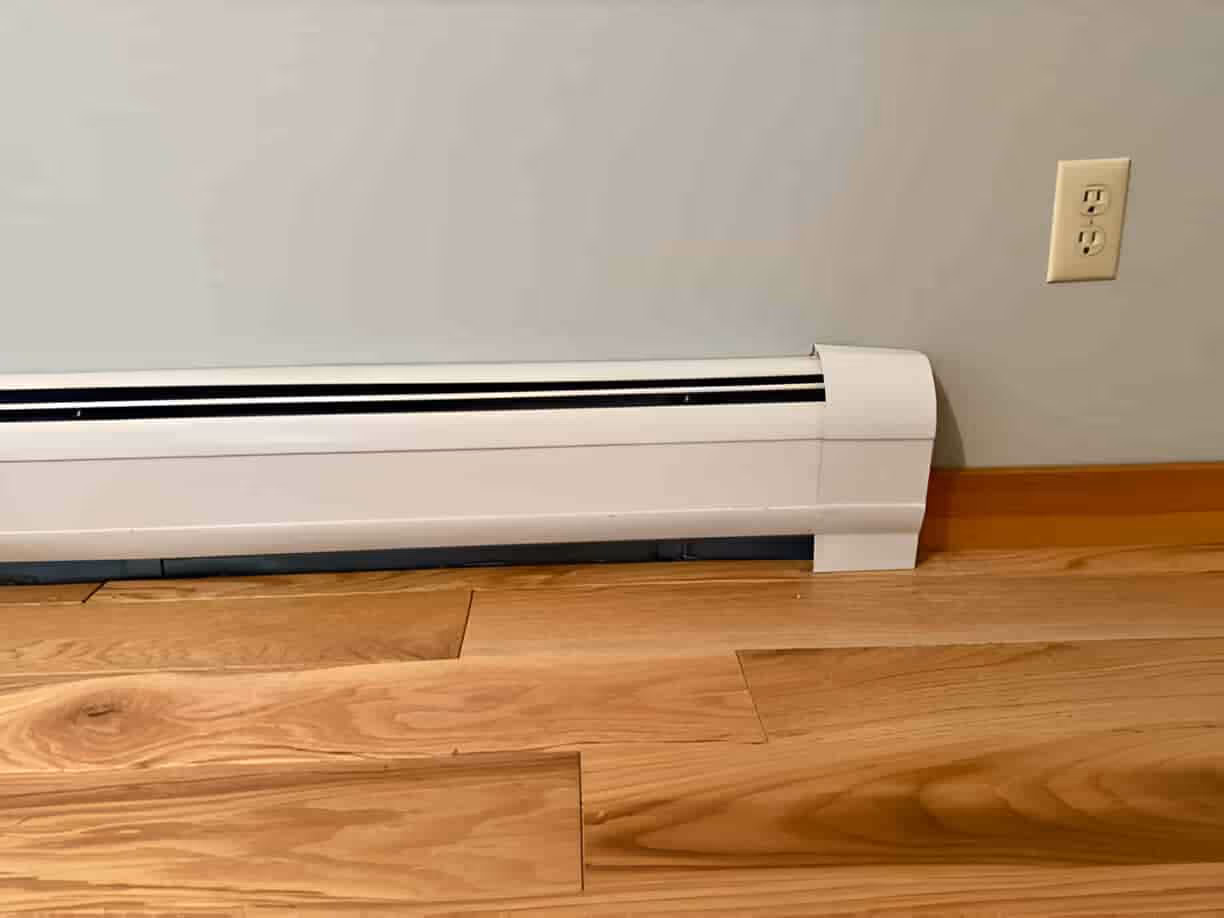 White electric baseboard heater mounted on wooden floor near wall outlet