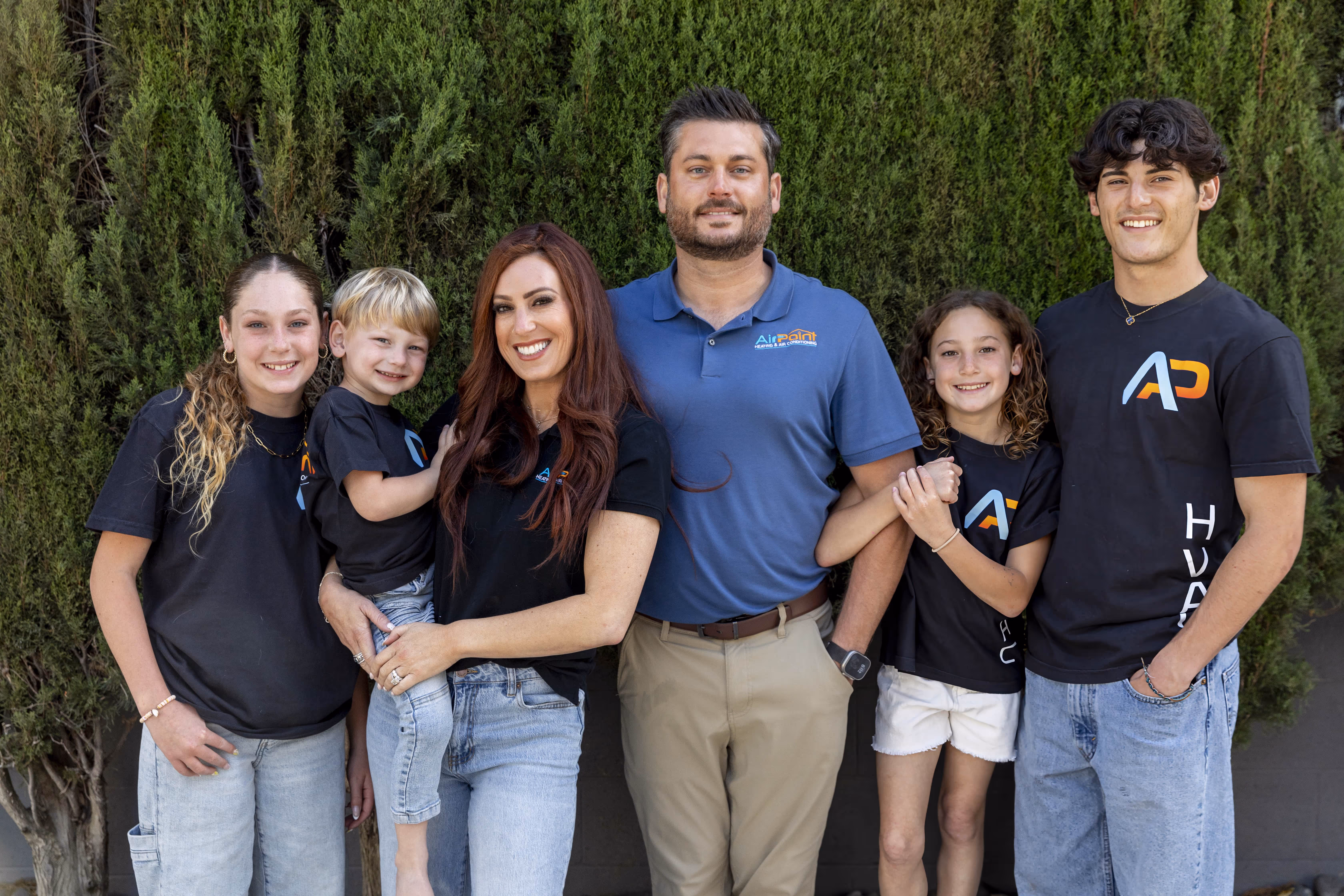 Family of six posing together in matching company shirts against green hedge