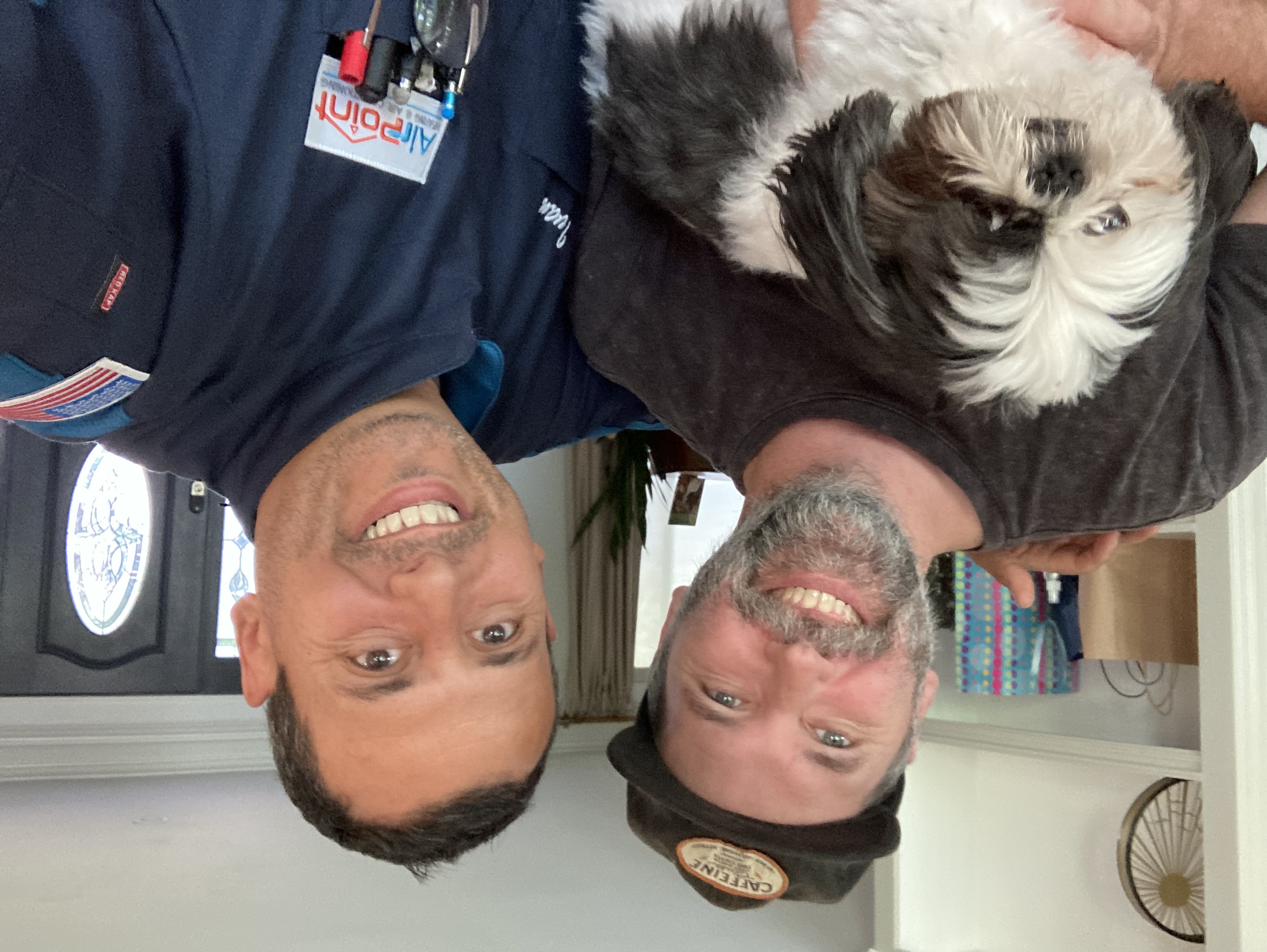 Two smiling men holding a black and white fluffy dog at home