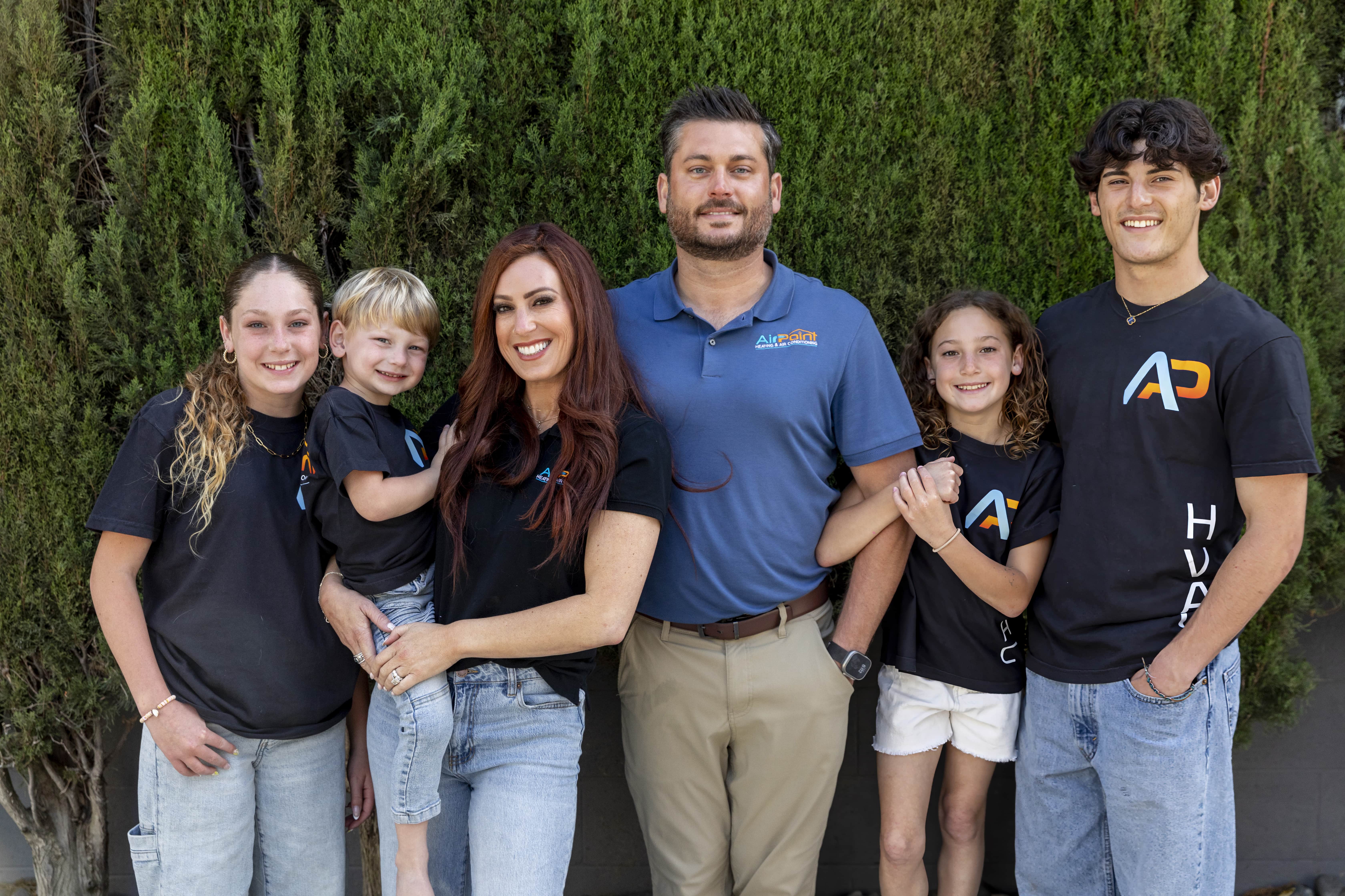 Family of six posing together in matching company shirts against green hedge