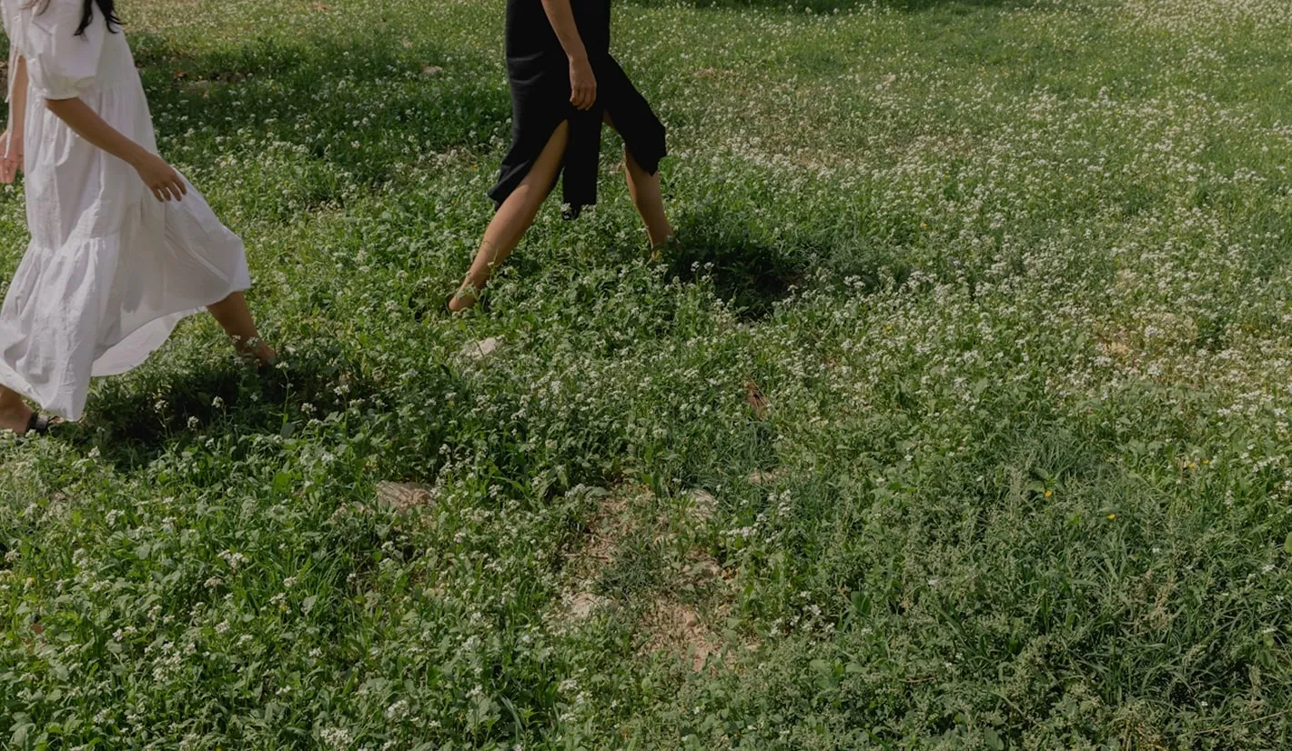 Two people wearing dresses walk through a grassy field with small white flowers. 
