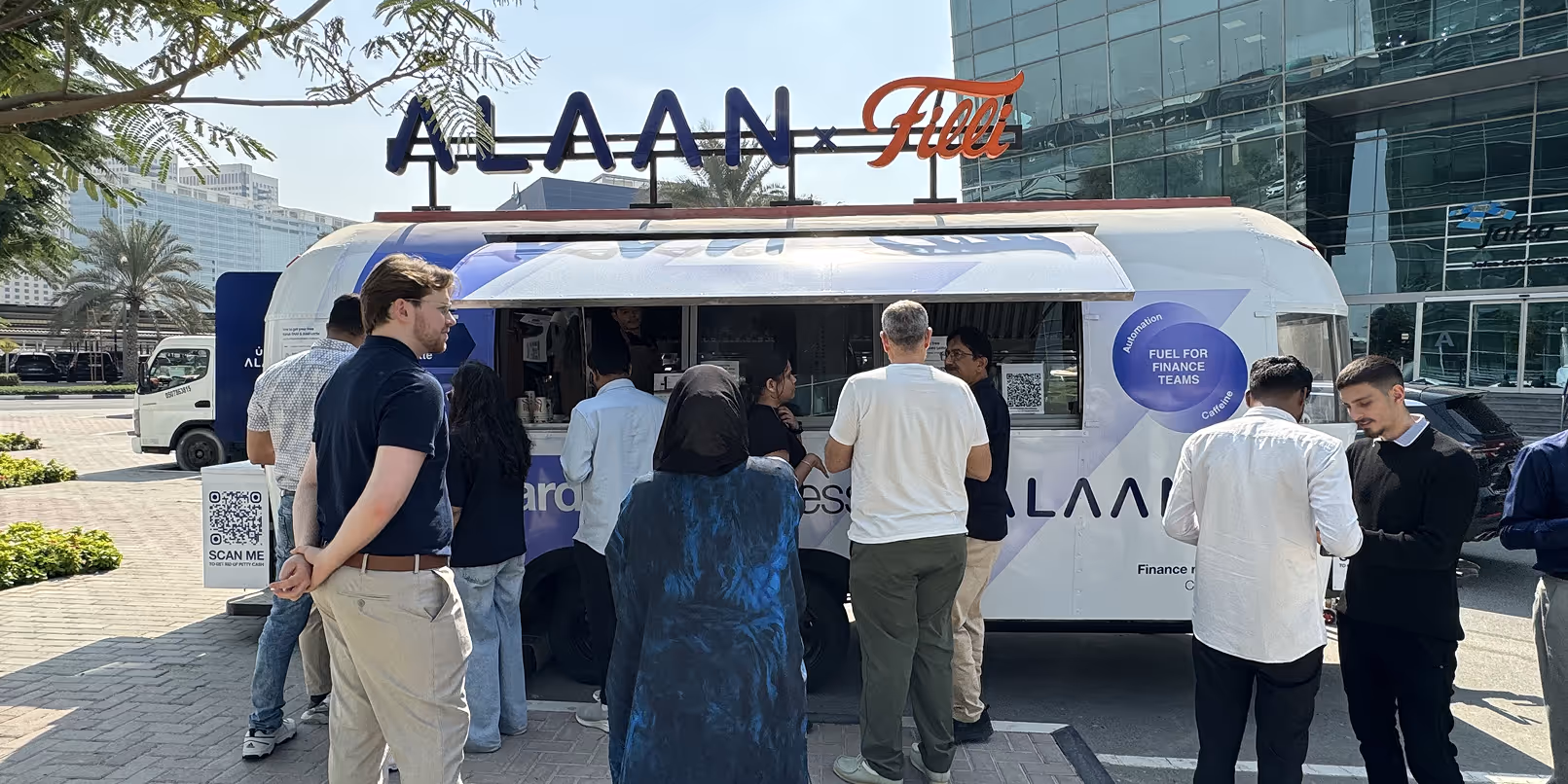 People standing in line outside a white and blue Alaan Filli coffee truck in an urban area with palm trees and modern buildings.