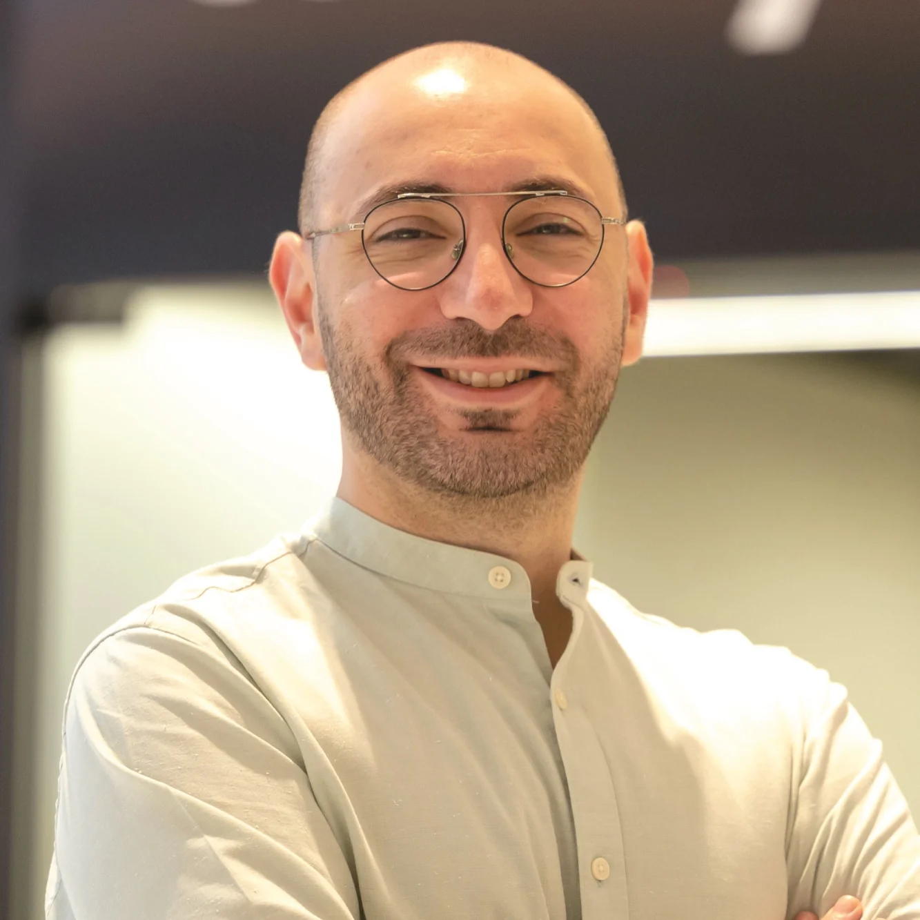 Smiling bald man with glasses wearing a light-colored buttoned shirt stands with arms crossed.