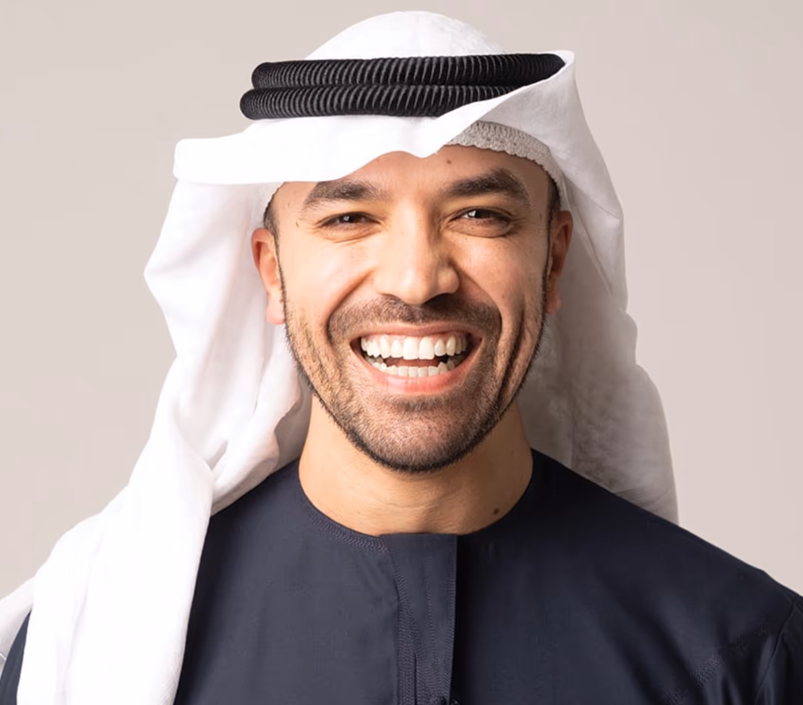 Smiling man wearing traditional white headscarf and dark garment on a beige background.
