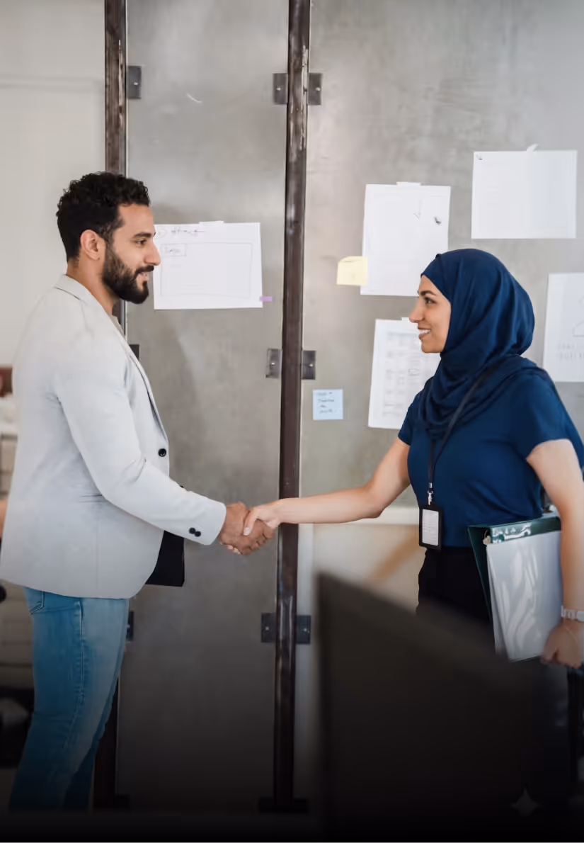 Two professionals, a man and a woman wearing a hijab, shaking hands in an office setting.