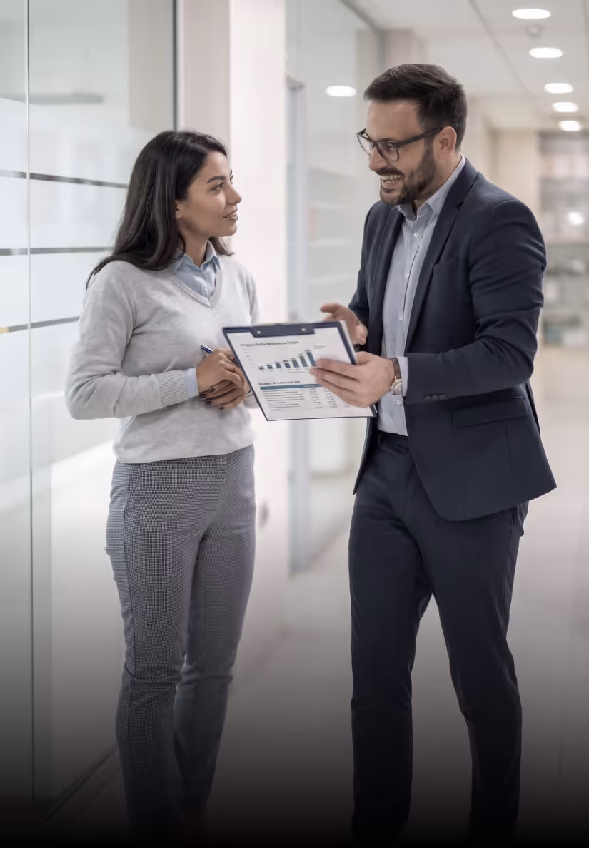 Two business professionals discussing a report with graphs in a bright office hallway.