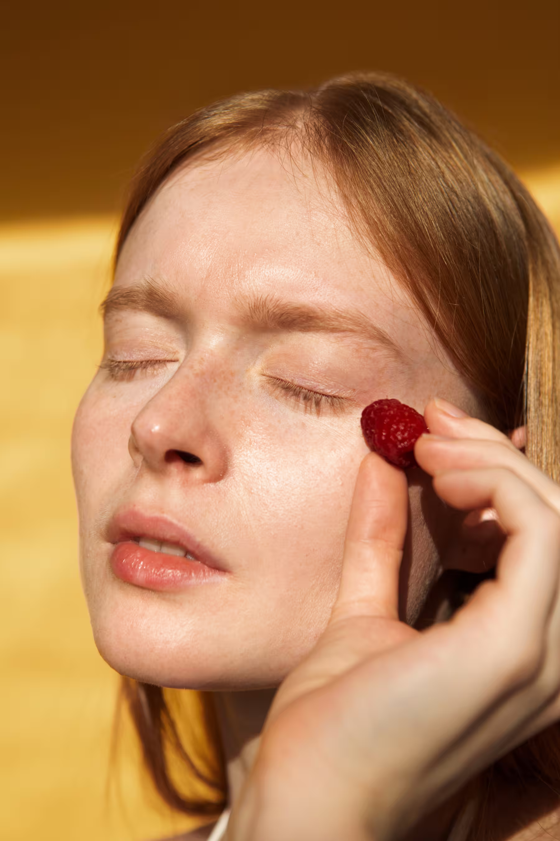 Femme aux cheveux roux tenant une framboise près de sa joue avec les yeux fermés.
