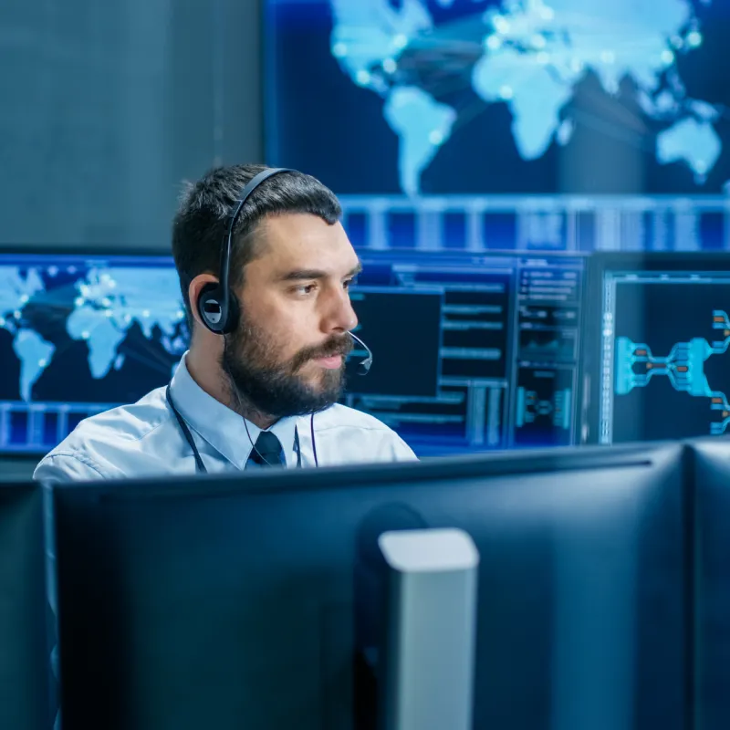 Man wearing headset and tie working at multiple computer monitors with world maps and data on screens.