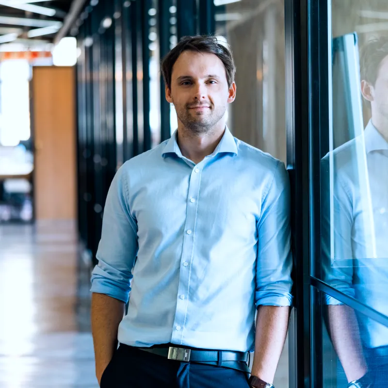 Smiling man in a light blue shirt standing with hands in pockets leaning against a glass wall in an office hallway.