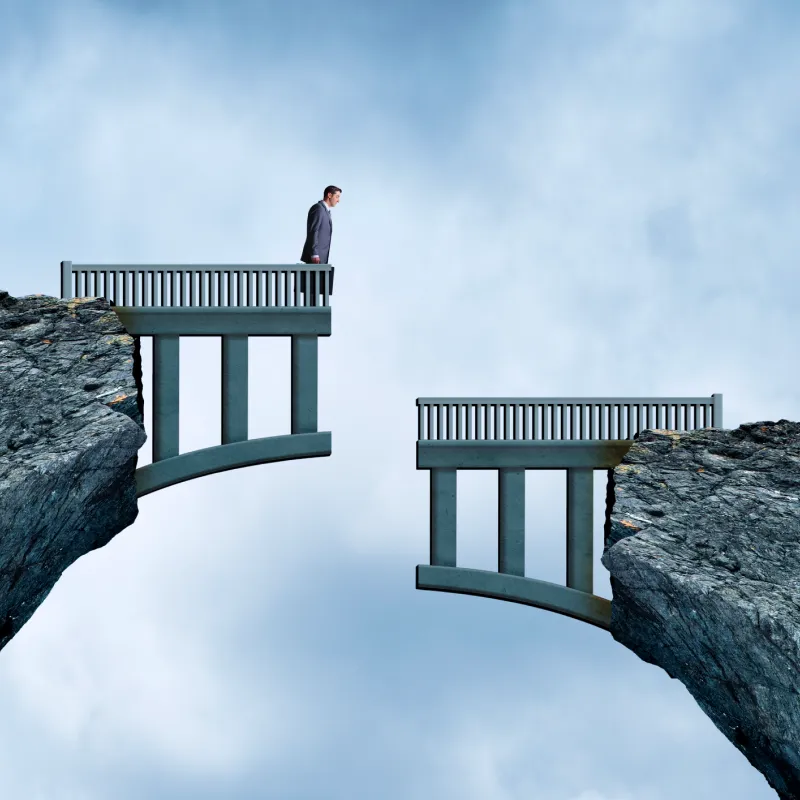 Man in business suit standing at the edge of a broken bridge between two cliffs.