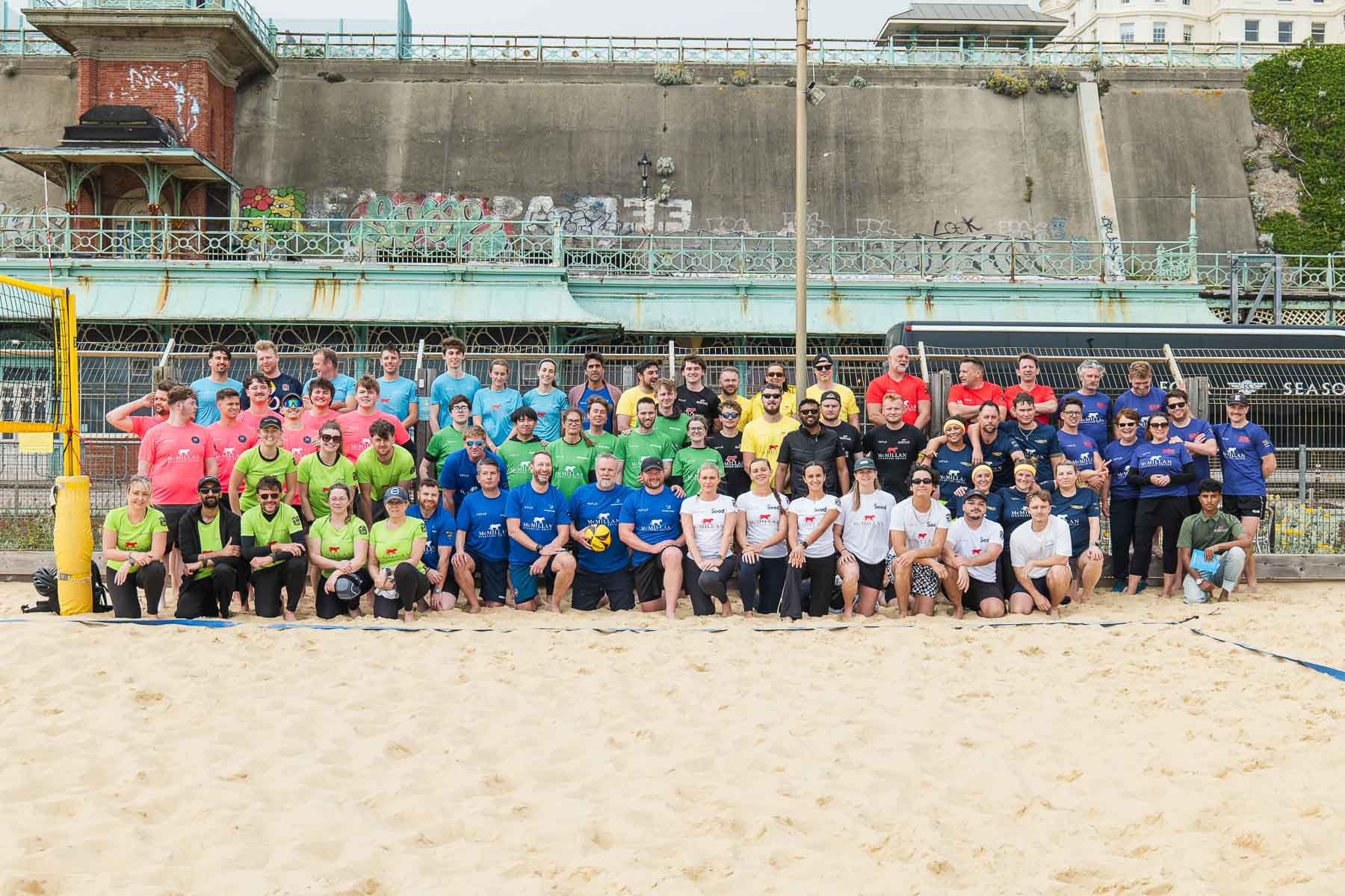 A large group of people wearing colorful shirts pose together on a sandy beach volleyball court.