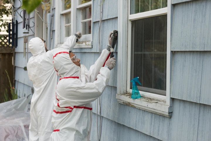 Two workers in protective suits perform maintenance on a blue wooden house, using tools near a window with a spray bottle nearby.