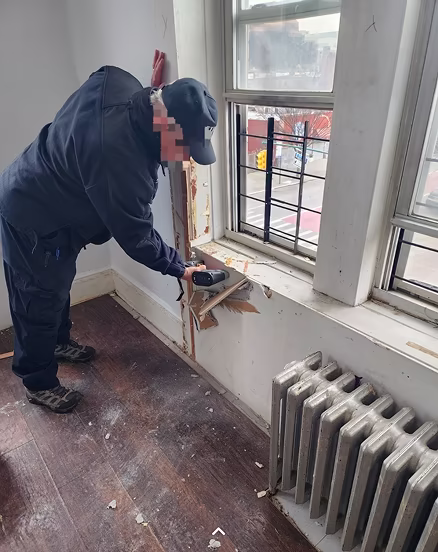 Person using a power tool to remove damaged wood around a window inside a room with a radiator and hardwood floor.