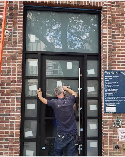 Man installing or cleaning a large glass door set in a brick building near a 'Work in Progress' sign.