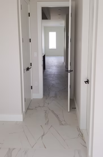 Narrow hallway with white walls and marble-patterned floor tiles, doors on both sides, leading to a room with a bright window.