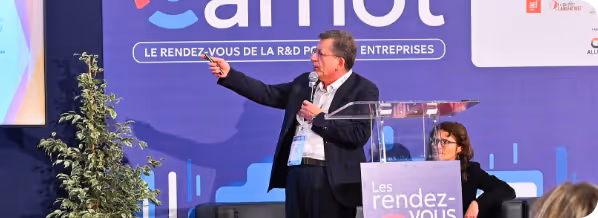Un homme en costume présente avec un micro et pointe un écran lors d'une conférence professionnelle.