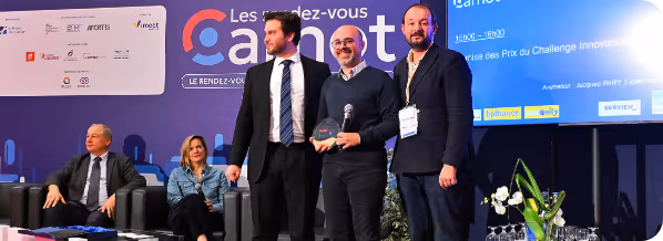 Trois hommes sur scène lors d'un événement, deux tenant un trophée et un micro, avec public assis en arrière-plan.