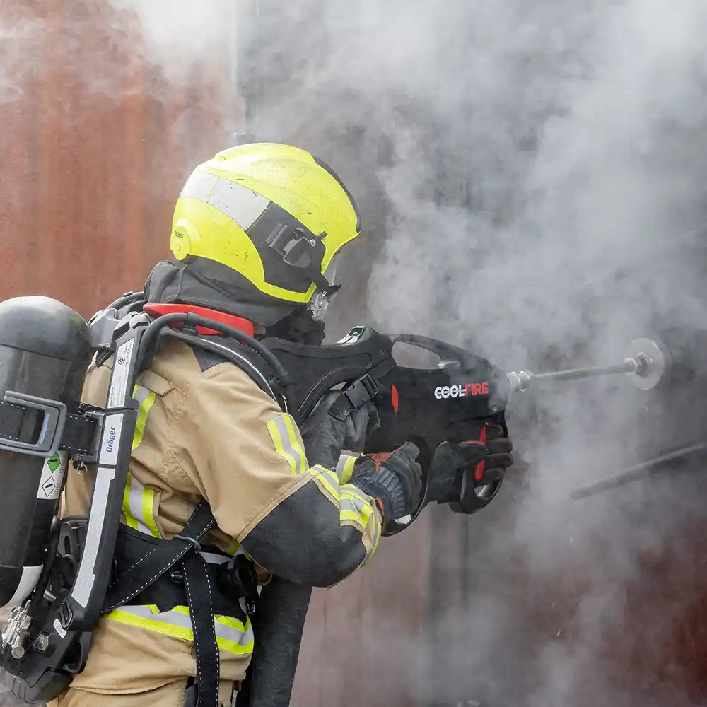 Brandweerman doorboort een stalen deur met de Coolfire-lans, om rookgassen van buitenaf te bestrijden. Dichte rookontwikkeling toont de intensiteit van de situatie. Coolfire maakt het mogelijk om veilig, effectief en zonder binnentreden een knockdown te geven.
