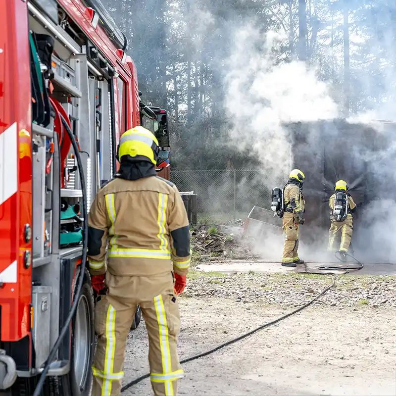 Brandweerlieden bestrijden rookontwikkeling met het Coolfire-systeem tijdens een training. De slanghaspel is aangesloten op het blusvoertuig, terwijl het team veilig en doeltreffend van buitenaf optreedt bij een gesimuleerde binnenbrand.