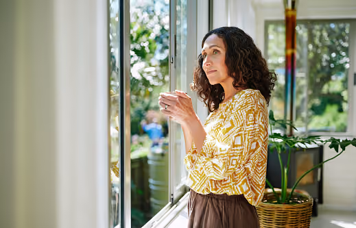 A woman in a yellow blouse holding a cup of coffee and looking out the window on a sunny day