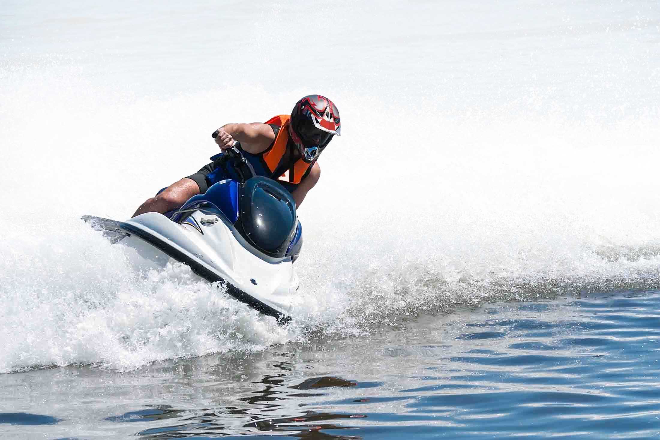 Person wearing a helmet and orange life jacket riding a blue and white jet ski on water, creating a large splash.