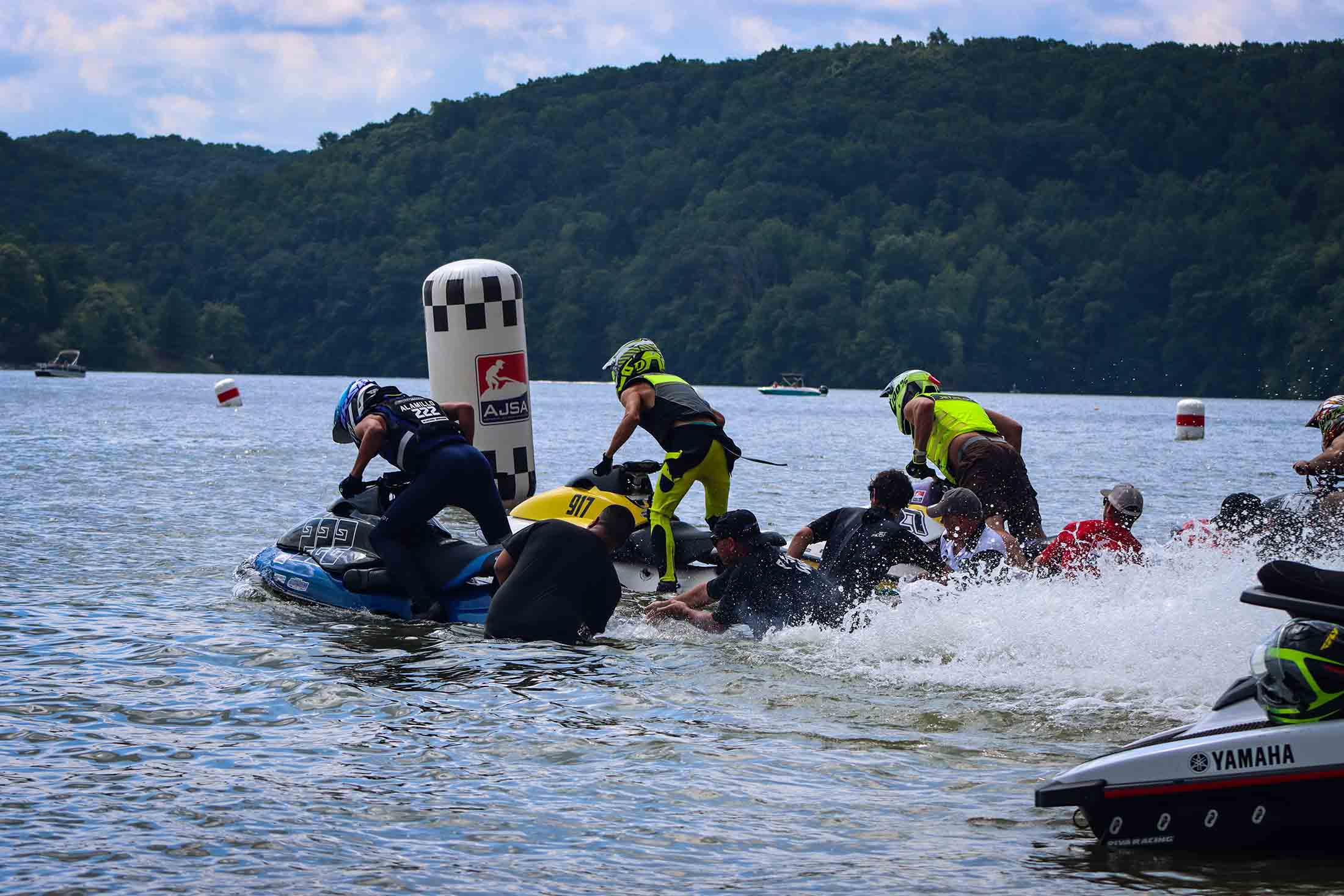 Jet ski racers pushing their watercraft into the water at a lakeside race start with forested hills in the background.