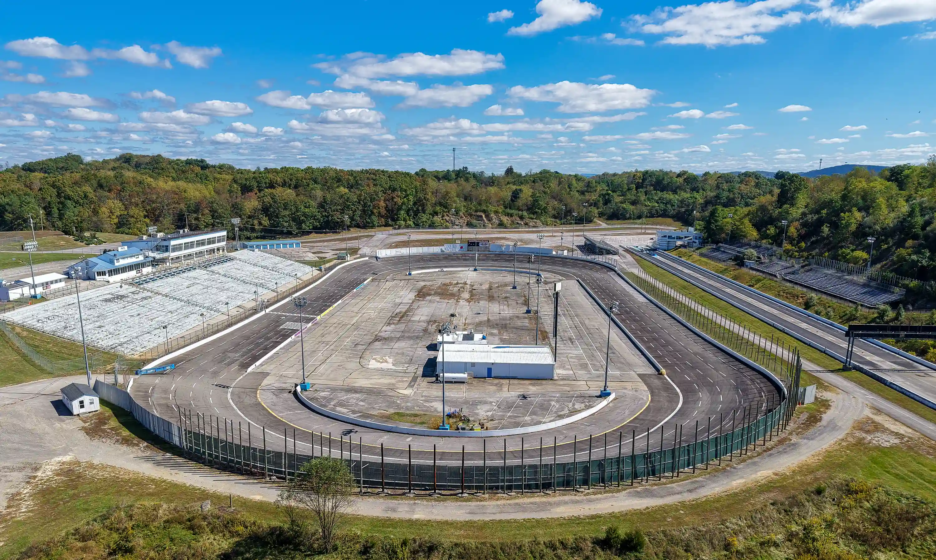 Empty oval race track surrounded by forest under a blue sky with scattered clouds.