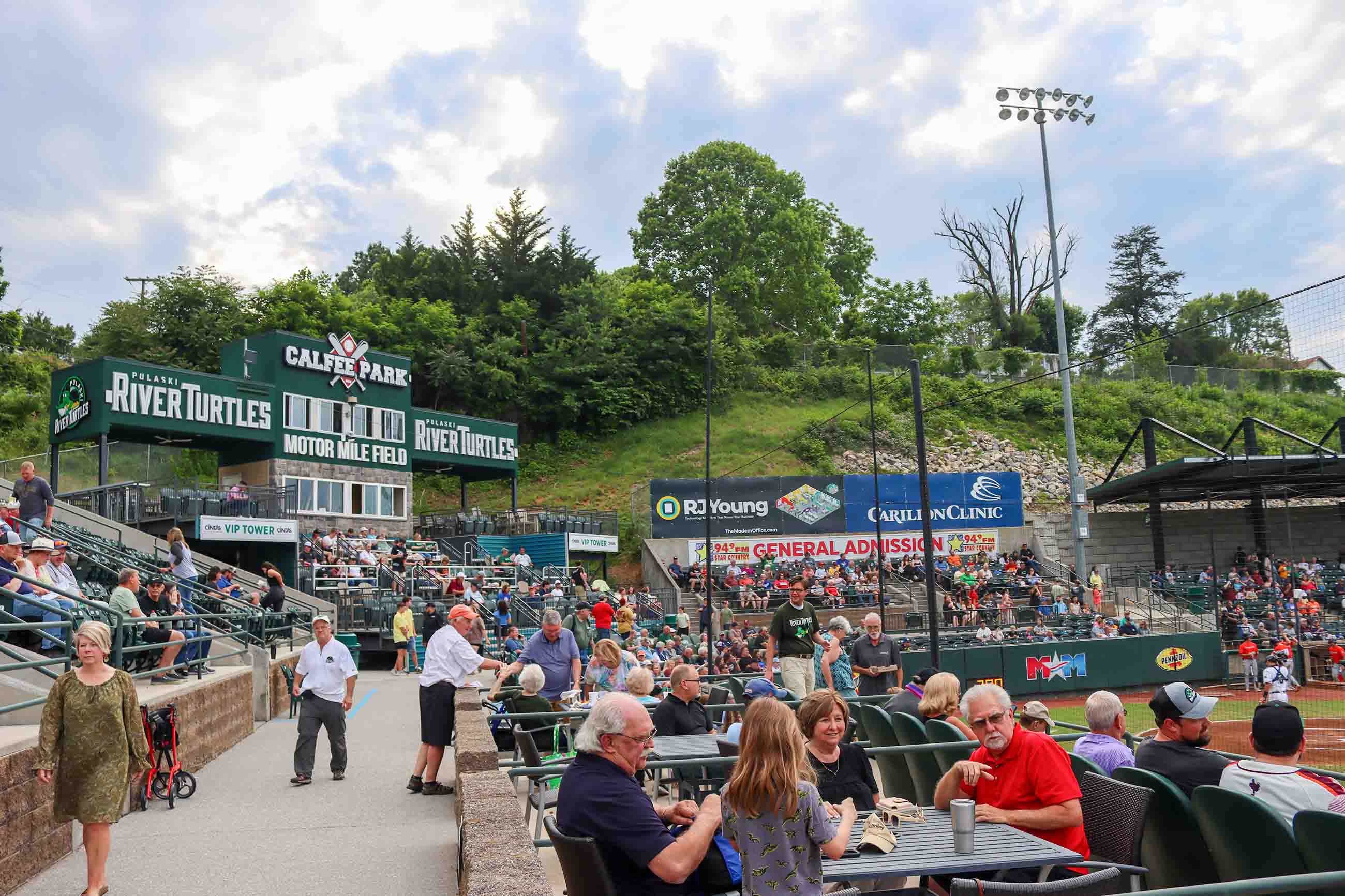 Crowd of people seated and walking near the stands at Calfee Park, home of the Pulaski River Turtles baseball team, on a partly cloudy day.