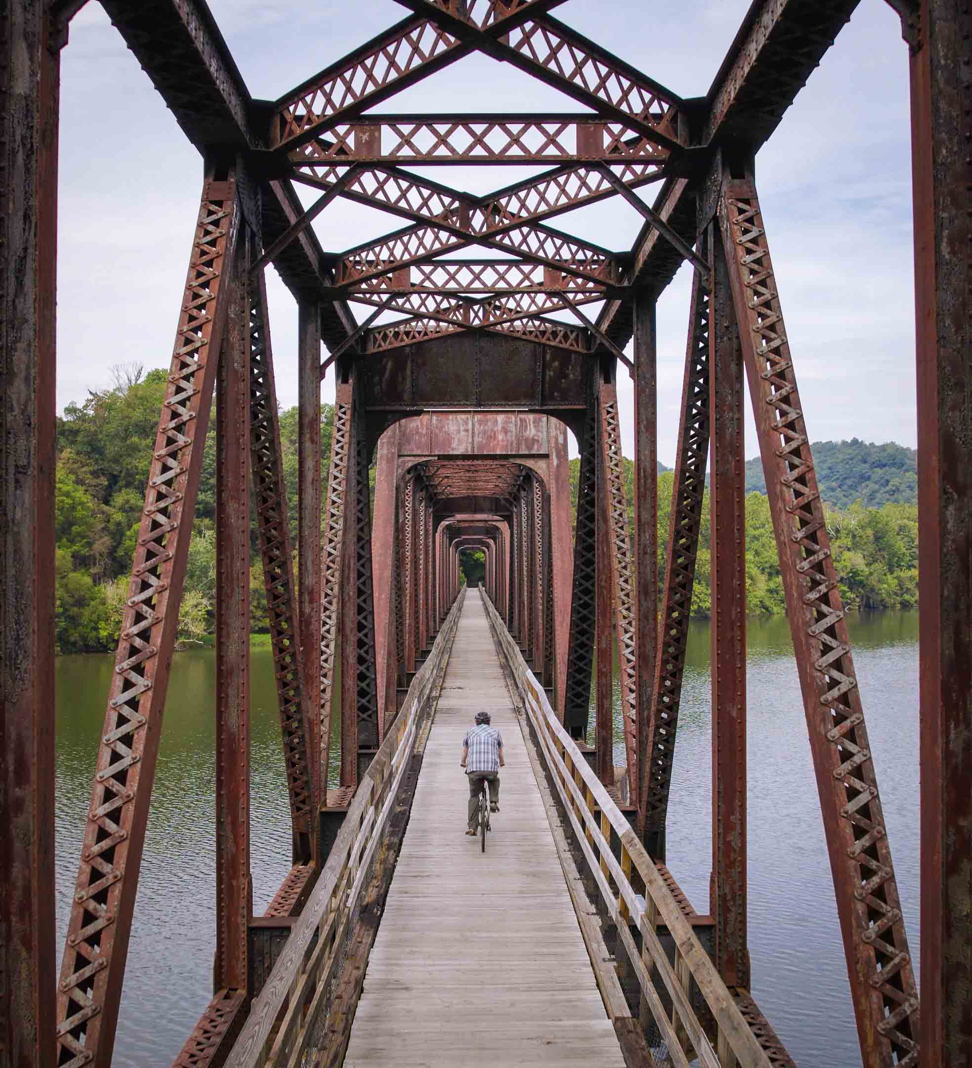 Person riding a bicycle on a wooden bridge with rusty metal trusses over a river surrounded by trees.