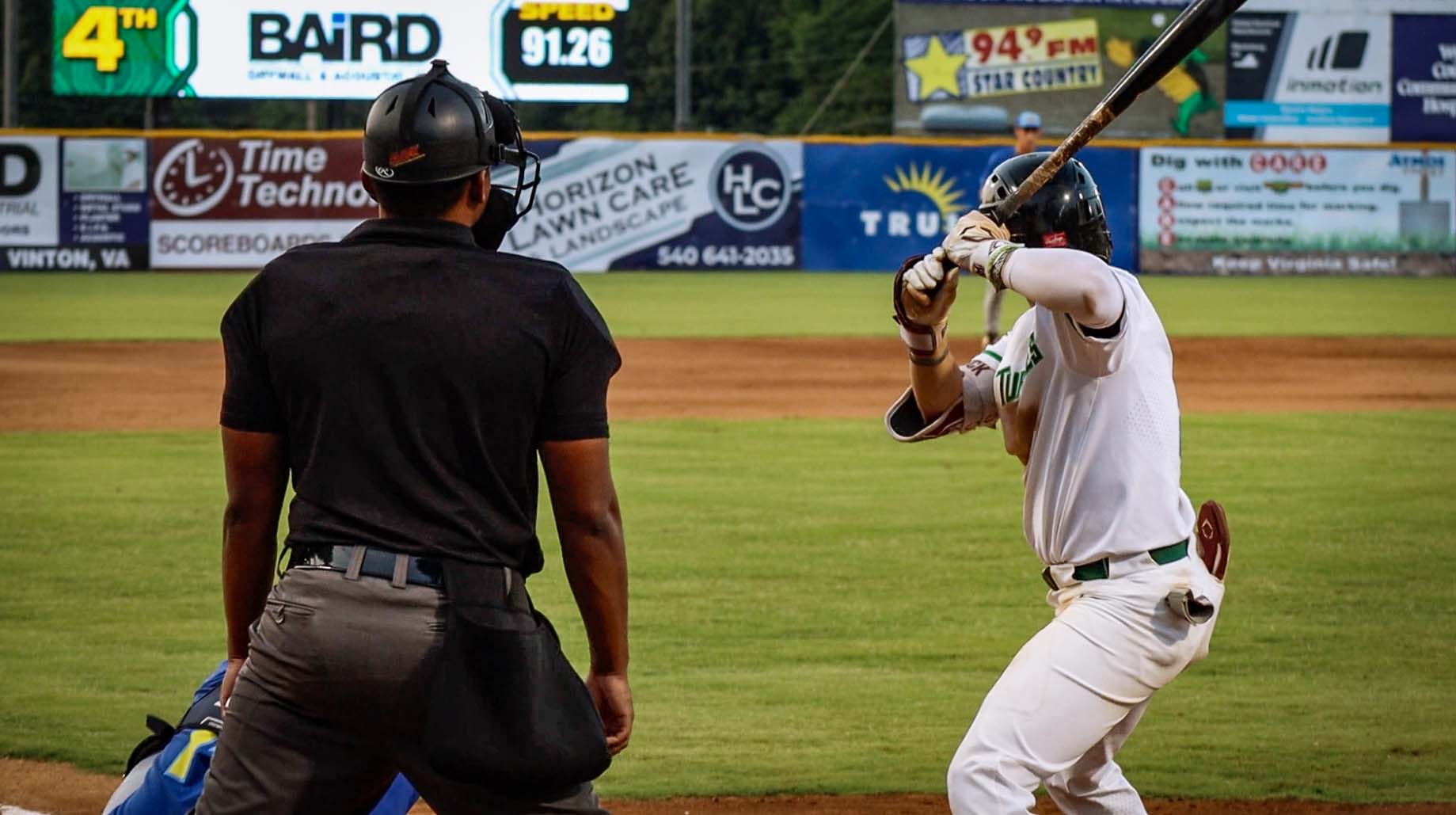 Baseball batter in white uniform preparing to swing as umpire in black shirt and helmet stands behind him on the field.
