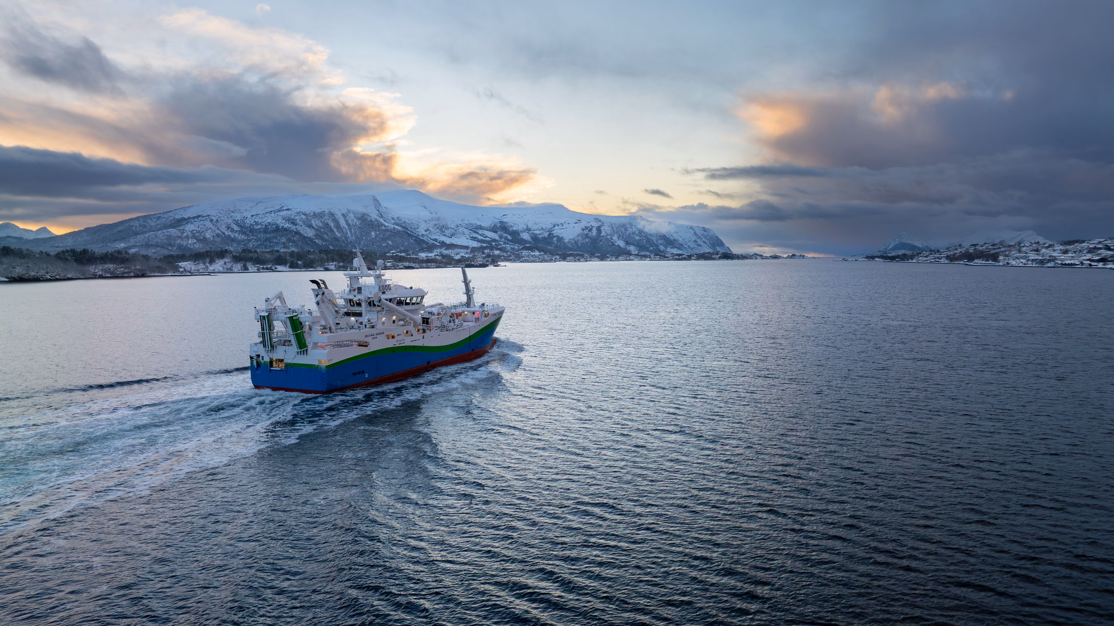 Fishing vessel with a blue, green, and white hull sailing on calm water with snow-covered mountains and a cloudy sky at sunset in the background.