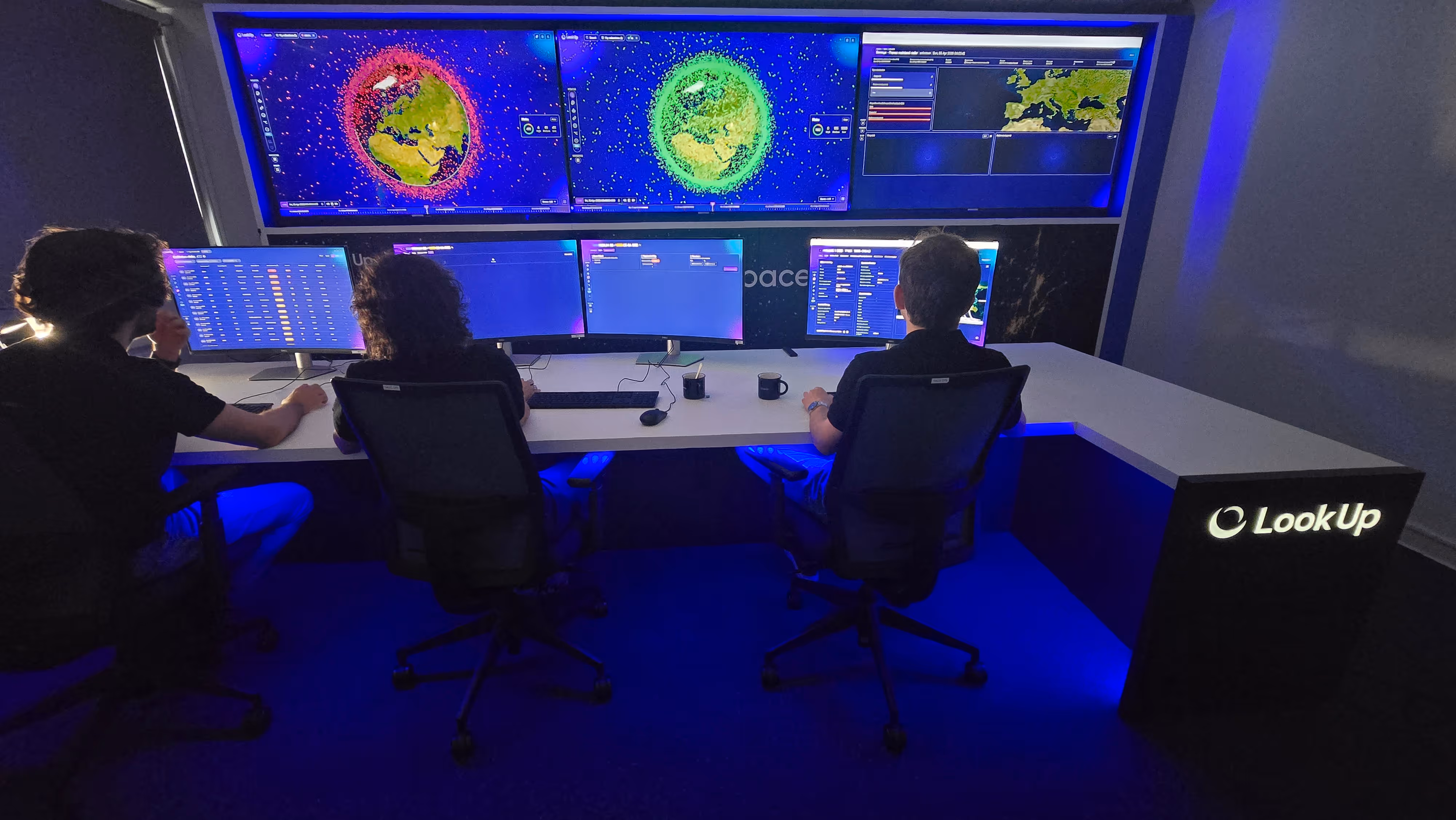 Three people seated at a control desk with multiple monitors showing Earth and data in a dimly lit room.