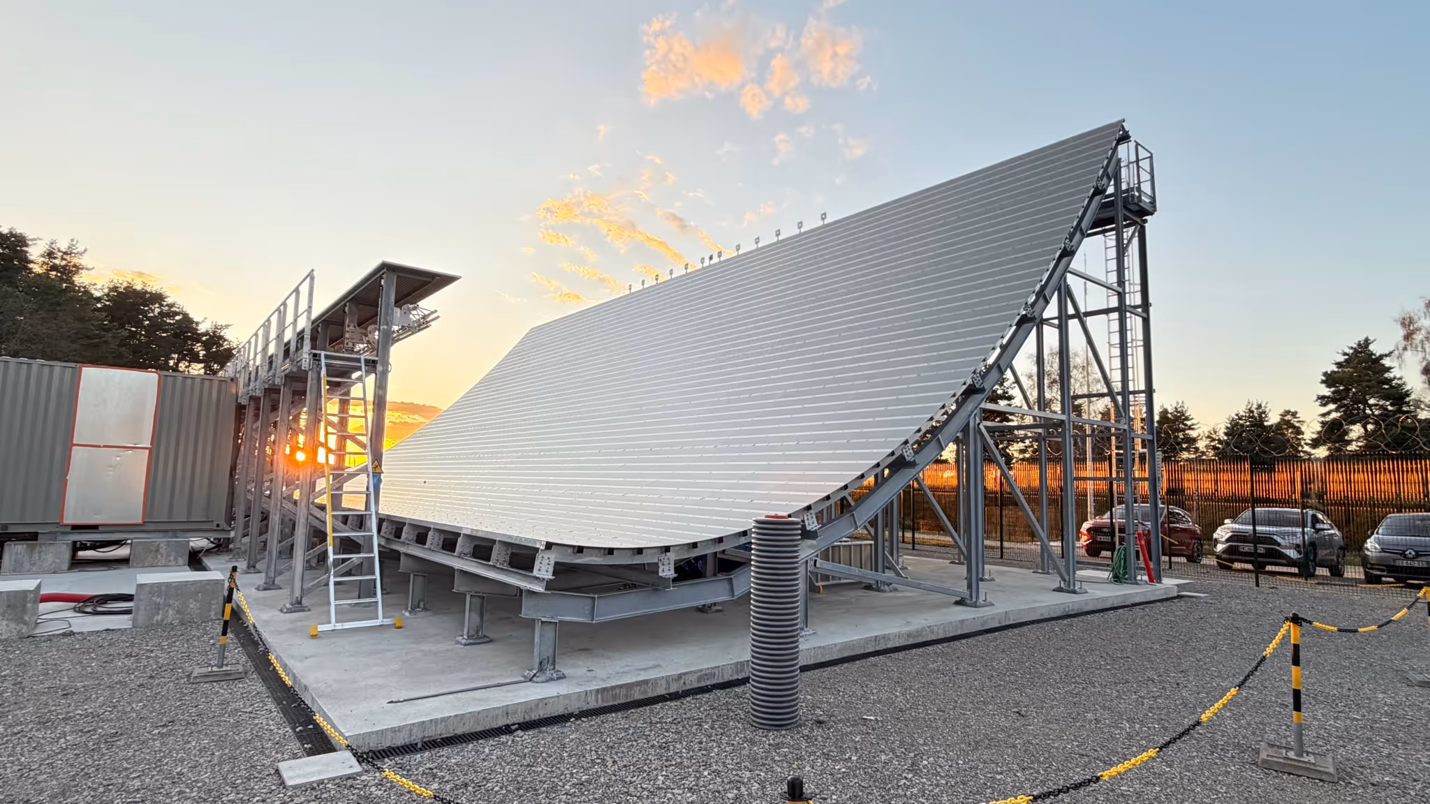 Large curved metal structure on a platform with cars and sunset in the background.