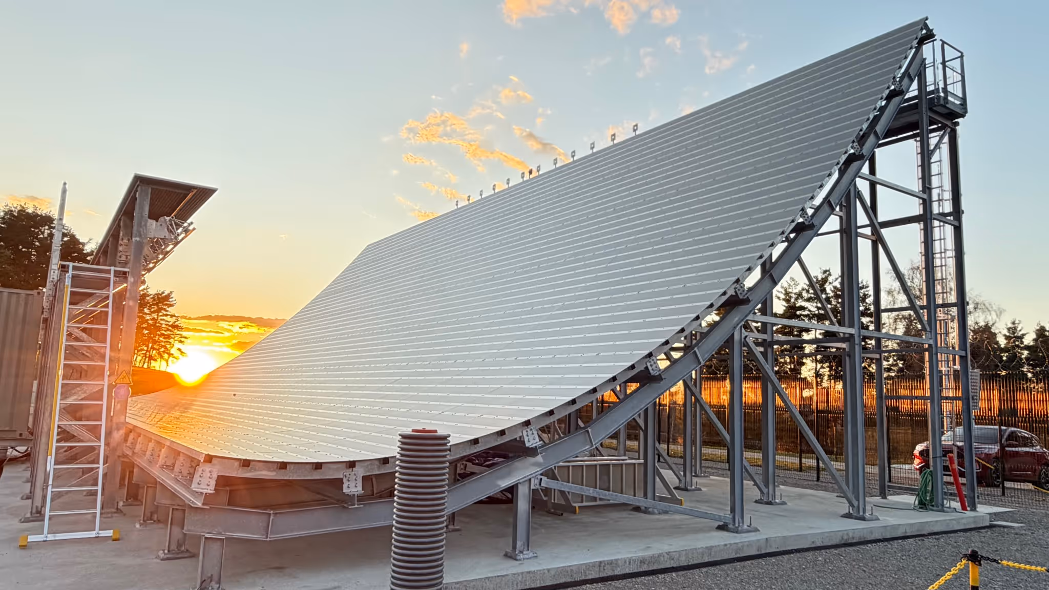 Large curved solar panel array on metal frame at sunset with trees and car in background.
