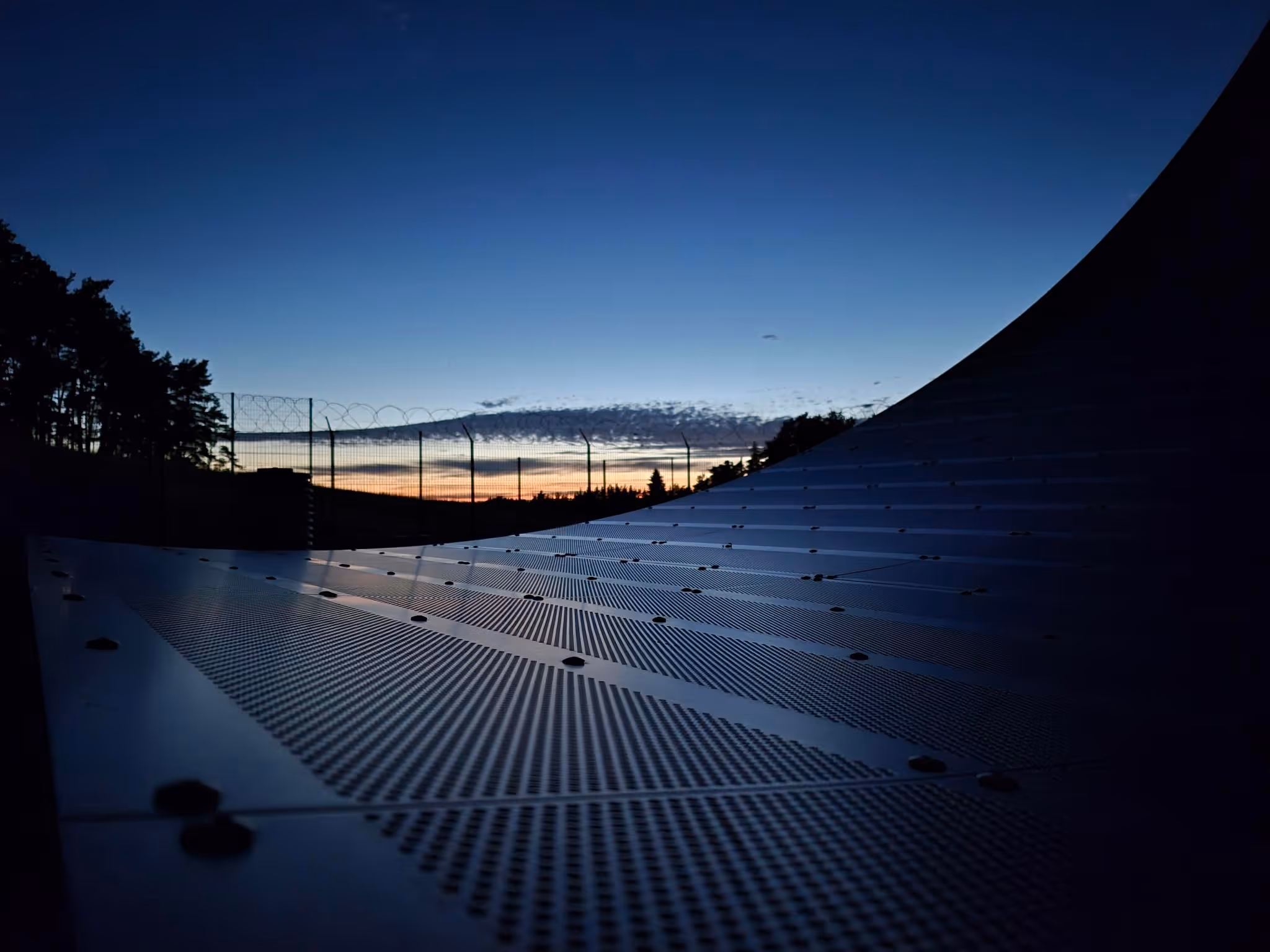 Curved solar panels reflecting a sunset with fence and trees in the background at dusk.