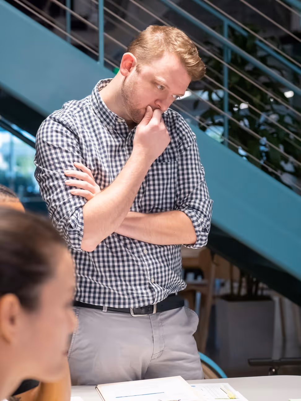 Man in checkered shirt standing with hand on chin in modern office interior, thinking.