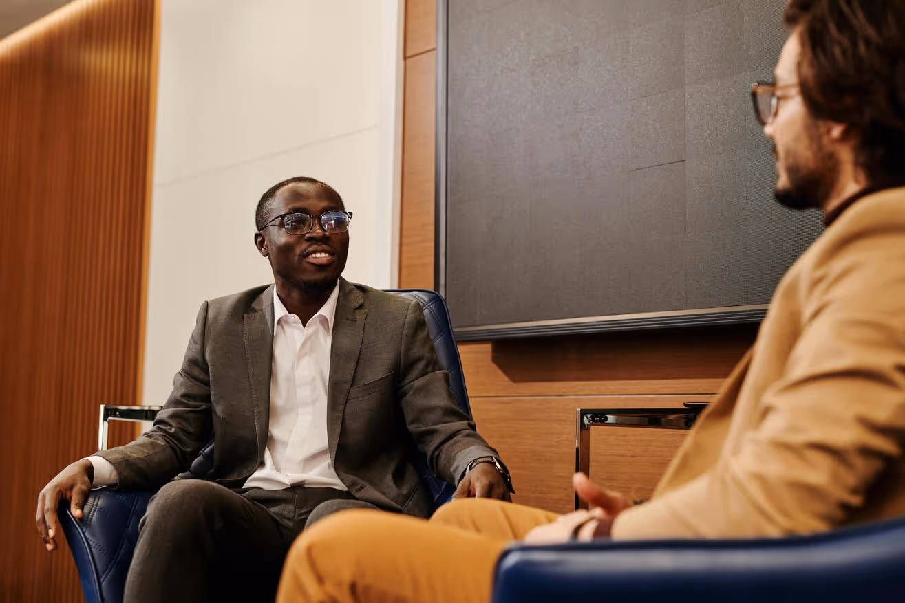 Two men in suits sitting and talking in an office with wood panel walls and modern chairs.