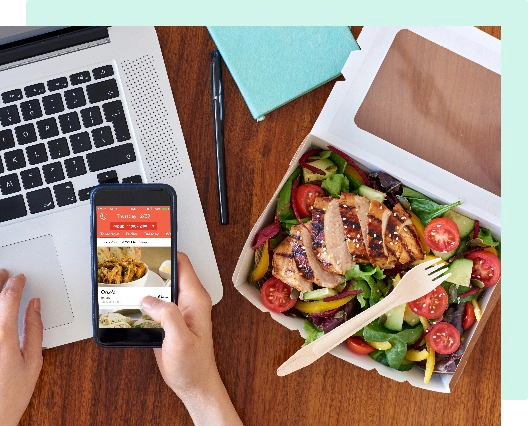 photo of a person lookin at the Fooda app on their phone with a delicious salad with grilled chicken beside them and a laptop in the background