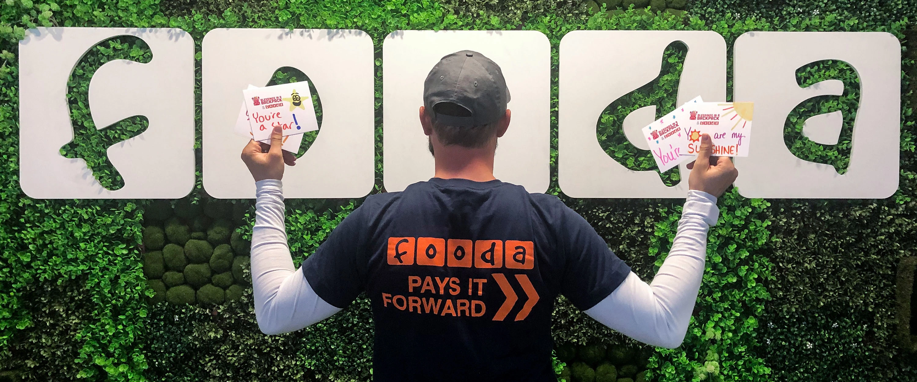 Fooda employee in a shirt that says 'Fooda Pays it Forward' standing in front of the Fooda logo on a green wall